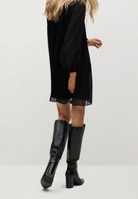 Vestido negro con plisados y mangas largas de tul, combinado con botas de cuero negras hasta la rodilla con tacones cuadrados y un acabado liso y elegante.