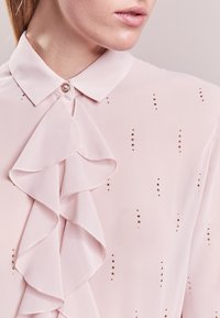 Helle rosa Bluse mit gerafftem Vorderbereich, einem Knopfkragen und metallischen Akzenten, die eine glatte Textur und zarte Nähte aufweist.