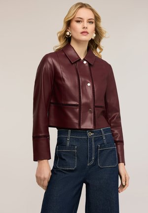 Blondynka nosząca złote kolczyki koła, bordową skórzaną krótką kurtkę z zatrzaskami oraz ciemnoniebieskie dżinsy z wysokim stanem i przednimi kieszeniami.