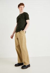 Tricou din bumbac verde închis, cu mâneci scurte, purtat cu pantaloni bej cu picioare largi și saboți negri cu detalii de cusătură galbenă.