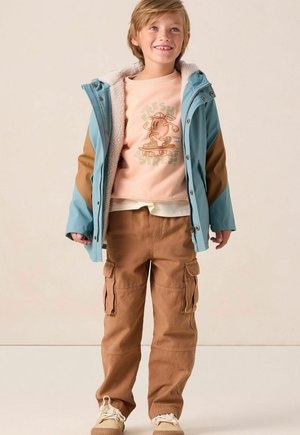 Veste d'extérieur bleue et marron avec doublure en polaire, sweat-shirt graphique beige, et pantalon cargo marron avec poches. Des baskets beiges complètent le look.