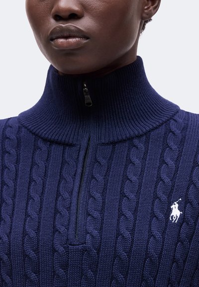 Gros plan d'une personne portant un pull en maille torsadée bleu marine avec une fermeture éclair mi-longue et un logo brodé représentant un joueur de polo blanc sur la poitrine.