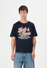 Mornarska pamučna majica s bijelim i crvenim natpisom "Jack & Jones" i "DENIM GOODS" u opuštenom kroju. Uparena s svijetloplavim trapericama.