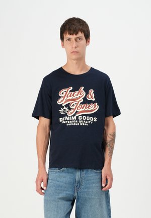Navy cotton t-shirt featuring white and red text "Jack & Jones" and "DENIM GOODS" with a relaxed fit. Paired with light blue denim jeans.