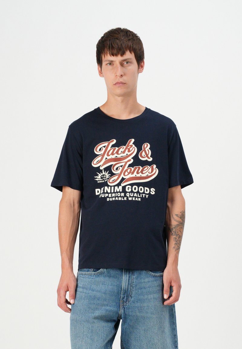 Mornarska pamučna majica s bijelim i crvenim natpisom "Jack & Jones" i "DENIM GOODS" u opuštenom kroju. Uparena s svijetloplavim trapericama.
