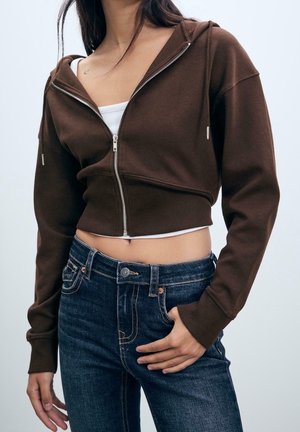 Femme portant un sweat à capuche court marron zippé sur un top blanc avec un jean en denim bleu, main posée sur la hanche, cheveux longs et foncés partiellement visibles.