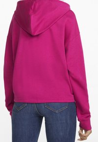 Sudadera fucsia con capucha ajustable, mangas raglán y un corte relajado, combinada con jeans de mezclilla oscuros con un diseño de bolsillo trasero en forma de V.