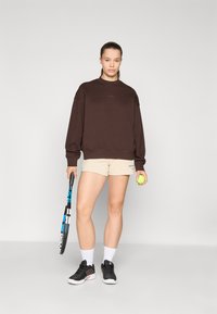 Sweatshirt oversize marron, shorts beiges, chaussures de sport noires, tenant une raquette de tennis et une balle de tennis, se tenant sur un fond neutre.