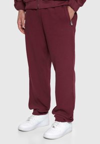 Bordeaux sweatpants med elastisk talje, sidelommer og subtil branding; parret med hvide sneakers med struktureret overflade.