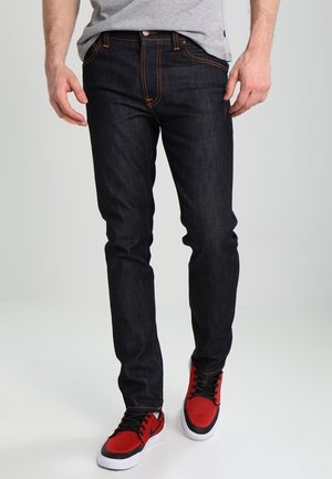 Homme portant un jean slim bleu foncé et des baskets rouges et noires, debout devant un fond gris clair.