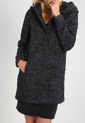 Femme portant un manteau à capuche texturé gris foncé avec des poches par-dessus une courte robe noire, une main dans la poche et l'autre touchant son visage.