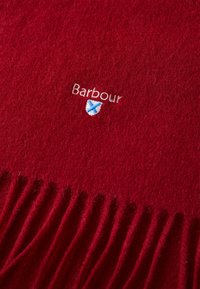 Écharpe en laine rouge avec des bords frangés. Présente un texte brodé "Barbour" et un petit logo en forme de bouclier bleu. Texture douce, apparence chaude.
