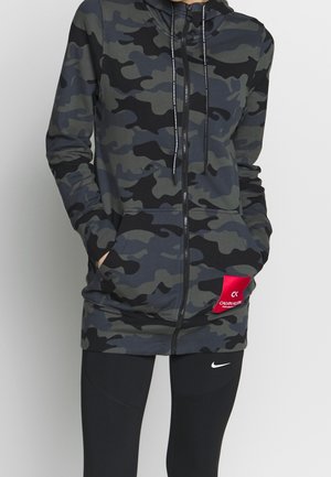 Persoon draagt een zwarte en donkergrijze camouflagehoodie met ritssluiting en een rode Calvin Klein-patch, gecombineerd met zwarte Nike-legging.