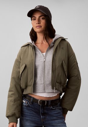 Frau trägt eine olivgrüne Bomberjacke über einem grauen Kapuzenpullover mit Reißverschluss, dunkler Mütze und blauen Jeans mit schwarzem Gürtel, steht vor einem schlichten Hintergrund.