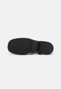 Must musta kingad musta kummiga, millel on tekstureeritud sooned ja muster haardevate omadustega. Siledad ääred ja kergelt tõstetud kanna disain. Minimalistlik bränding nähtav.