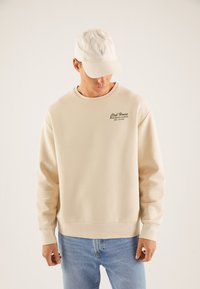 Beige genser med rund hals og ribbestrikkede mansjetter og kant, med grønn tekst. Kombinert med en lys farget baseballcap med logo broderi.