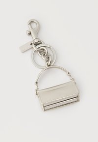 Μπρελόκ από ασημένιο μέταλλο με ένα μικρό γούρι σε σχήμα τσάντας και μια κλιπ, συνδεδεμένα με δύο στρογγυλούς κρίκους σε λευκό φόντο.