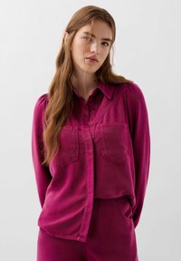 Camicia fucsia a maniche lunghe in tessuto morbido con una chiusura a bottoni frontale, due tasche sul petto e delicati plissè sulle spalle. Texture liscia, vestibilità strutturata.