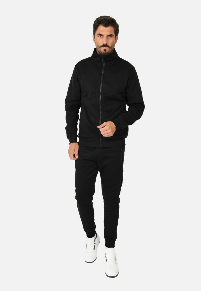 Giacca nera con zip e pantaloni coordinati in un tessuto liscio, abbinati a sneakers bianche. Caratterizzata da un colletto alto e un design slim-fit.