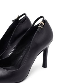 Zapatos de tacón alto de cuero negro con una textura suave, laterales abiertos y un pequeño detalle de hebilla en la correa del tacón.