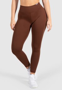 Bruine high-waisted leggings van rekbare stof, met een soepele textuur en naden, gecombineerd met witte sportschoenen.