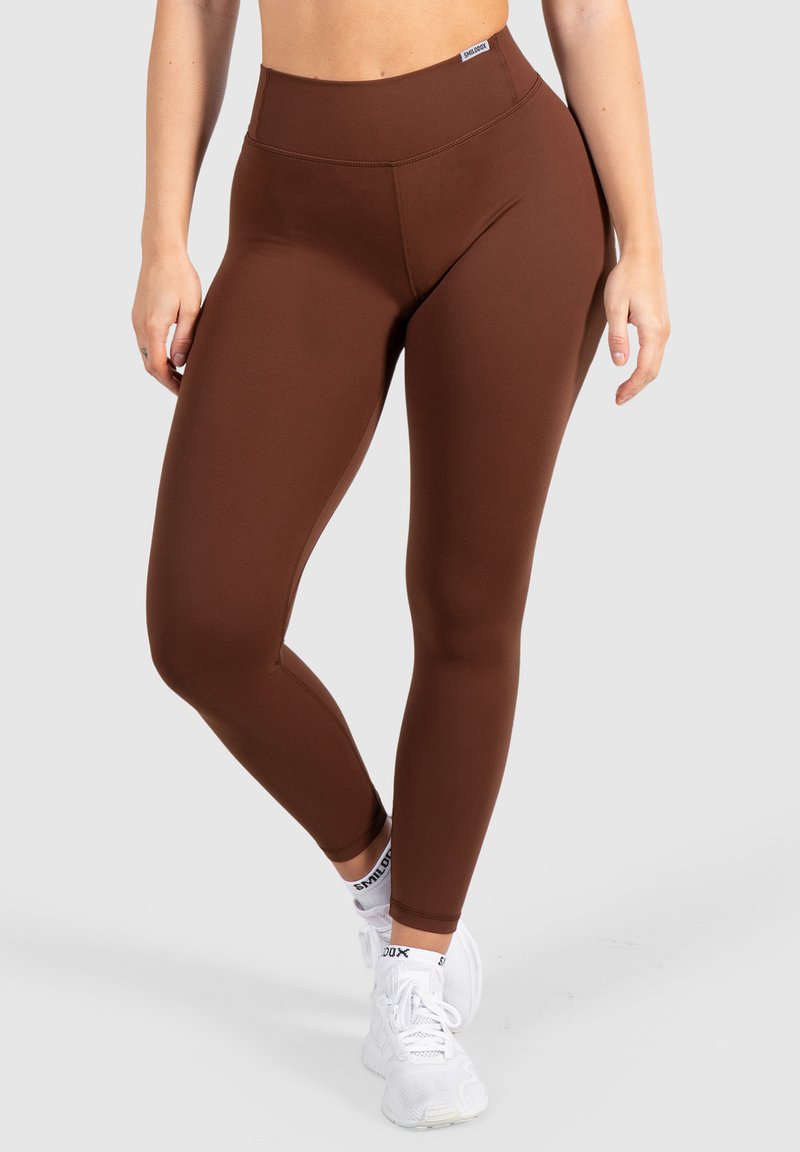 Bruine high-waisted leggings van rekbare stof, met een soepele textuur en naden, gecombineerd met witte sportschoenen.