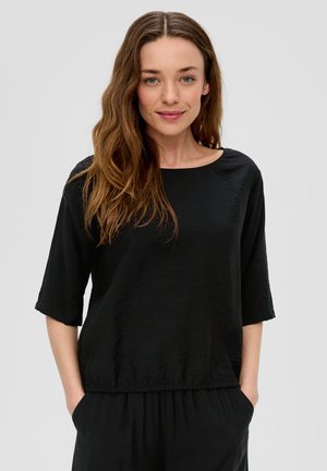 Zwarte top met korte mouwen, een brede halslijn en een licht gathered zoom. Gelaagd stof, casual ontwerp. Model heeft de handen in de zakken.
