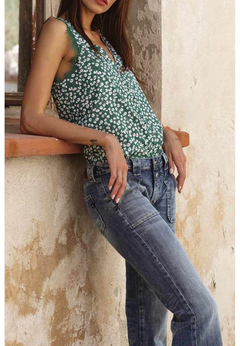Femme appuyée contre le bord d’un mur texturé, portant un haut sans manches vert à fleurs et un jean bleu délavé, les mains posées sur les hanches.
