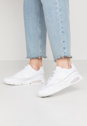 Ben iført lyseblå korte jeans med frynsete kanter og hvide atletiksko med mesh og syntetiske detaljer.