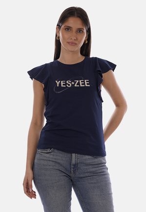 Donna con lunghi capelli scuri che indossa orecchini a cerchio dorati, top blu navy con maniche arricciate e la scritta "YES ZEE", e jeans azzurri, in piedi con la mano sui fianchi.