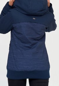 Mörkblå jacka med huva, med en texturerad övre panel och en randig nederdel. Ribbad mudd och nederkant, logopatch på ryggen.