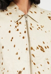 Μπεζ μπουφάν από συνθετική γούνα με καφέ πουά μοτίβο, διαθέτει γιακά που διπλώνει και κεντρικό μεταλλικό φερμουάρ για κλείσιμο.