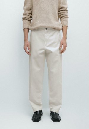 Persona che indossa un maglione beige lavorato a maglia, pantaloni beige chiaro e scarpe slip-on in pelle nera, in piedi davanti a uno sfondo semplice.