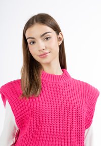 Felle roze gebreide mouwloze trui met een hoge hals, een textuurpatroon en brede schouderopeningen, gedragen over een witte langemouwen shirt.