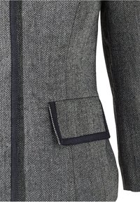 Saka na míru s herringbone vzorem v kombinaci černé a šedé barvy. Má strukturovaný tvar, texturovanou látku a kapsu s kontrastním lemováním.