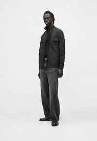 Black lightweight jacket with two front pockets, high collar, worn over a black shirt, paired with grey wide-leg pants and black shoes.