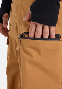 Braune wasserdichte Hose mit glatter Textur, ausgestattet mit einer Reißverschlusstasche am Oberschenkel und einem Zugband für einen einfachen Zugriff.