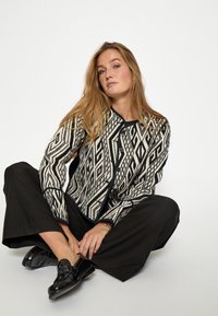 Czarna i biała kurtka w wzory z geometrycznymi deseniami, zapinana na guziki, z okrągłym dekoltem, zestawiona z czarnymi spodniami szerokimi w nogawkach i błyszczącymi mokasynami.