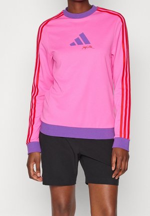 Osoba nosząca różową bluzę Adidas z czerwonymi paskami na rękawach i fioletowymi mankietami, połączoną z czarnymi szortami na jednolitym tle.