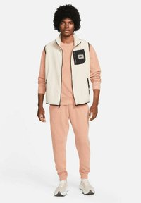 Beige bezrukávový svetr s černými kapsami, růžová dlouhá top s dlouhým rukávem a odpovídající tepláky. Sportovní boty s bílými a černými akcenty.