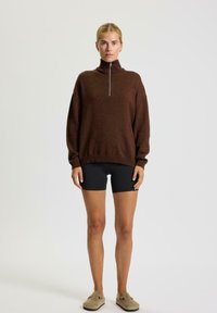 Brązowy sweter dzianinowy z wysokim kołnierzem i przednim zamkiem błyskawicznym, z długimi rękawami i luźnym krojem, zestawiony z czarnymi szortami i fakturowanym obuwiem.