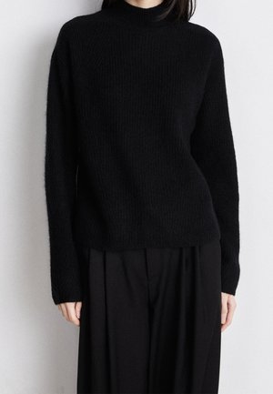 Osoba ubrana w czarny, długi, dopasowany sweter z wysokim kołnierzem oraz luźne czarne spodnie, stojąca na tle jednolitego, jasnego tła.