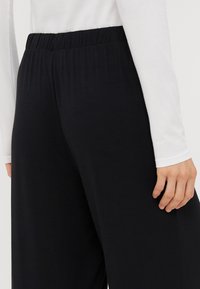 Μαύρο παντελόνι σε φαρδιά γραμμή με ελαστική μέση, κατασκευασμένο από μαλακό ύφασμα. Το ορατό χέρι ακουμπάει στον μηρό, με ένα λευκό μπλουζάκι μακρύ μανίκι.