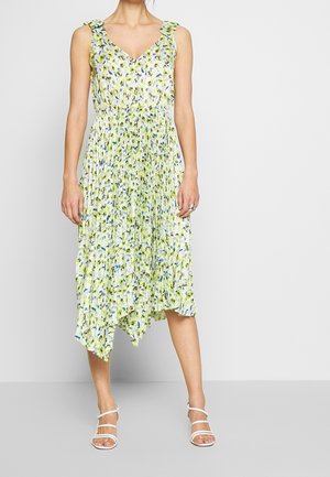 Frau trägt ein ärmelloses, plissiertes Midikleid mit gelbem und blauem Blumenmuster, Rüschenträgern und weißen Sandalen mit Riemchen.