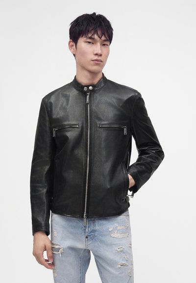 Jeune homme portant une veste en cuir noire avec des poches zippées et un jean bleu clair déchiré sur un fond blanc uni.