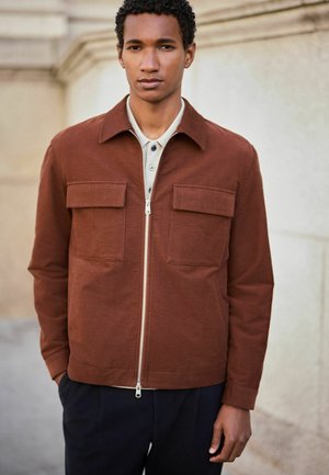 Young man wearing a rust-colored zip-up jacket with large front pockets over a light-colored shirt, standing against a beige stone wall.