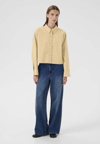 Żółta krótka bluzka z falbanami z przodu, długimi rękawami i spiczastym kołnierzykiem, w połączeniu z szerokimi niebieskimi dżinsami i czarnymi butami.