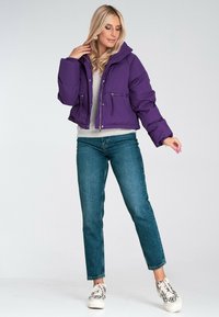 Fioletowa krótka kurtka puchowa z kapturem, z dwoma kieszeniami zapinanymi na zamek z przodu. Nosi się ją na jasnoszarym swetrze i w zestawieniu z niebieskimi dżinsami.