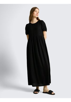 Frau trägt ein schwarzes, knöchellanges, kurzärmeliges Kleid und schwarze Slide-Sandalen, steht mit den Händen hinter dem Rücken und schaut zur Seite.