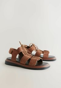 Las sandalias de ante marrón presentan múltiples correas anchas, un diseño de punta abierta y un detalle de cordones. La suela es negra con un acento marrón contrastante.